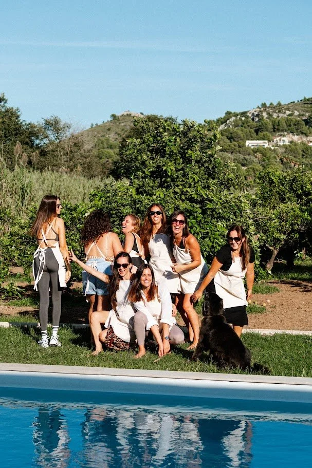 Grupo de mujeres en un jardín con árboles y hills en el fondo, algunas con delantal, junto a una piscina, disfrutando del día.