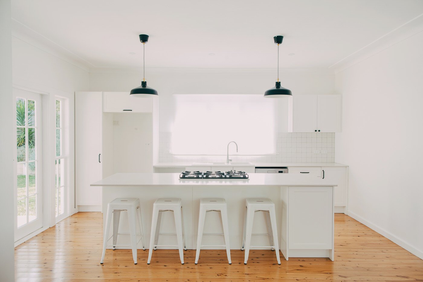Bright, minimalist kitchen with white cabinets, island with four white stools, wooden floor, black pendant lights, and large window letting in natural light.
