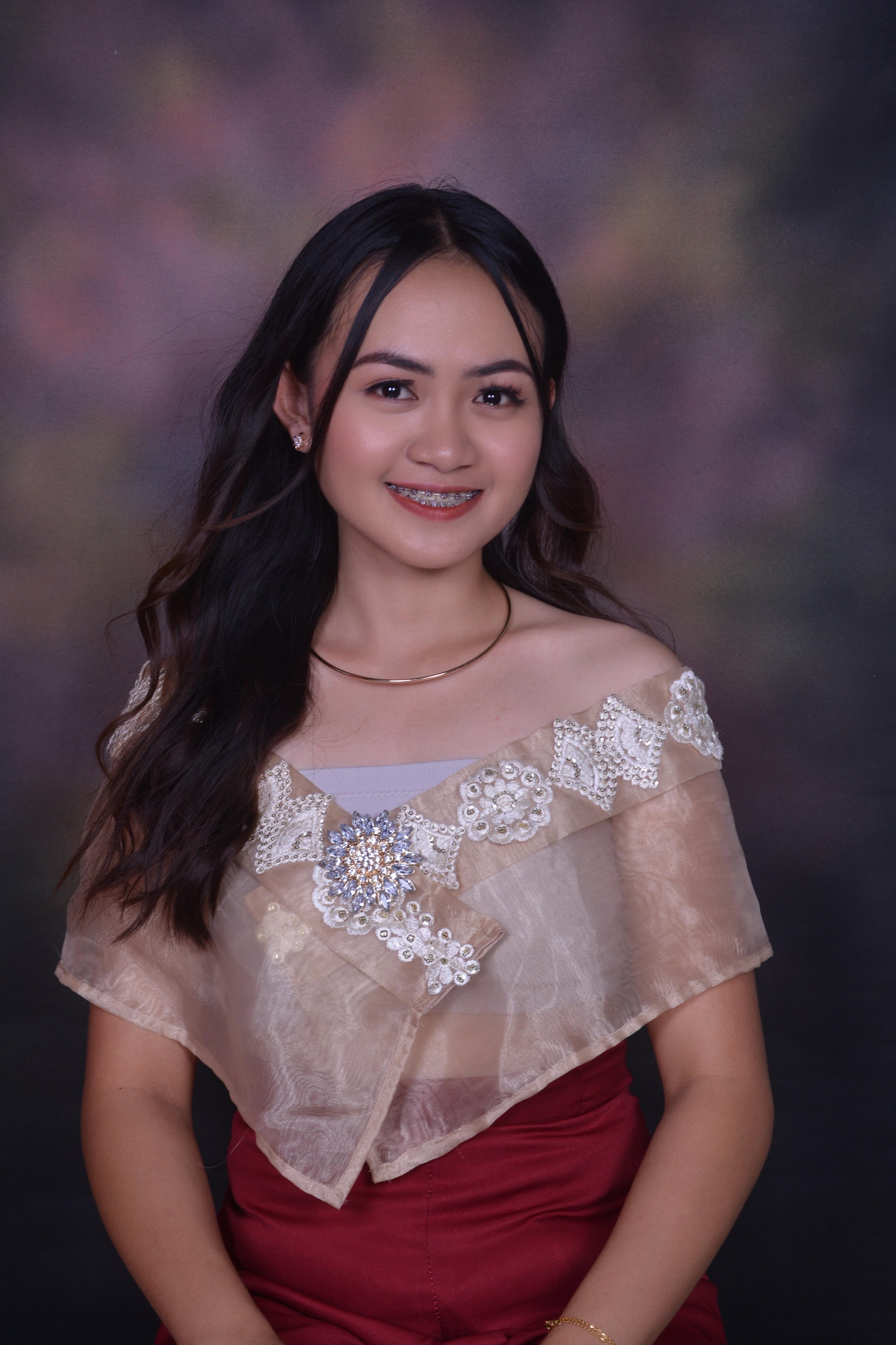 Young woman with long dark hair and braces, wearing an off-shoulder beige top with lace and pearl embellishments, a decorative brooch, and a red skirt, smiling against a blurred multicolored background.