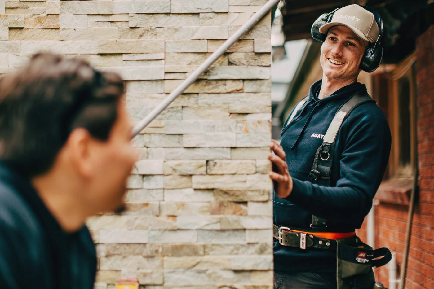 Builder in safety earmuffs smiling beside a stacked-stone wall detail, delivering Healthy Homes in Northern Beaches with careful finishes.