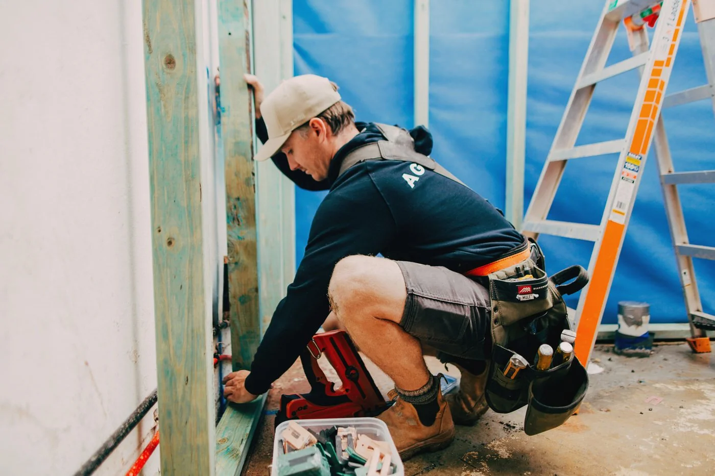 Builder kneeling to fix wall framing with tools and ladder on site, supporting Healthy Homes in Northern Beaches through quality workmanship.