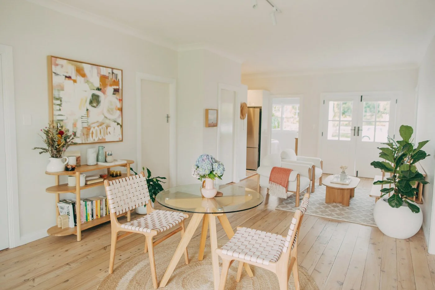 A generously proportioned open-plan interior with honey-toned hardwood floors, a glass round dining table, woven timber seating, and fresh blue hydrangeas in the foreground, the kind of everyday warmth that fills healthy homes in Dee Why