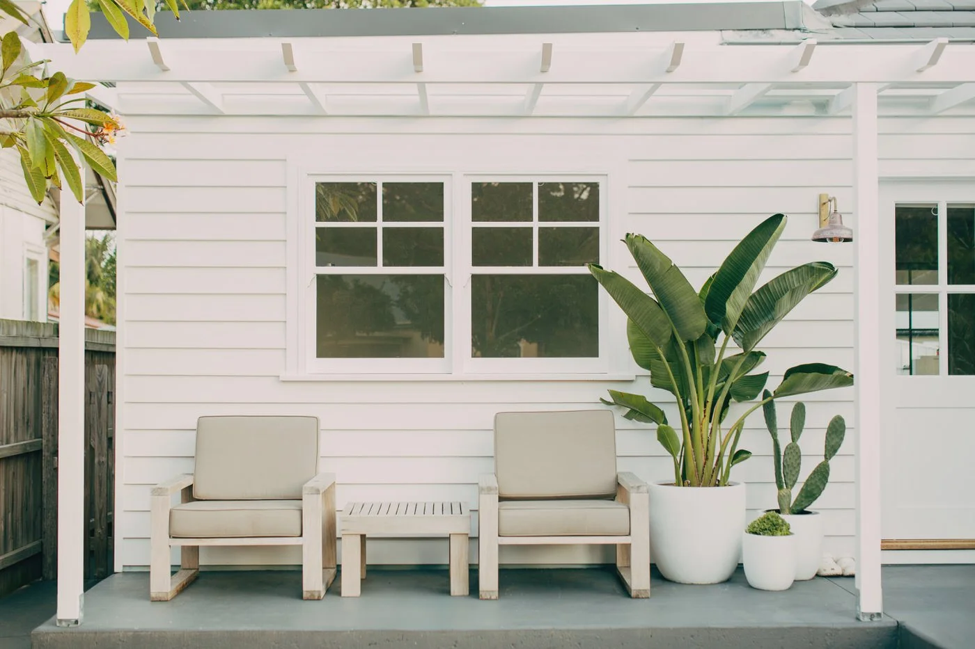 Two linen-cushioned timber chairs positioned beneath a clean white pergola with oversized ceramic planters and lush tropical foliage against a freshly painted weatherboard wall, a composed outdoor space that reflects healthy homes in Dee Why
