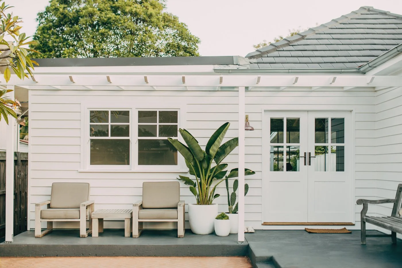 A thoughtfully renovated weatherboard exterior with a white timber pergola, French doors, and large leafy potted plants adding life to the covered outdoor area, the kind of quality craftsmanship behind healthy homes in Dee Why