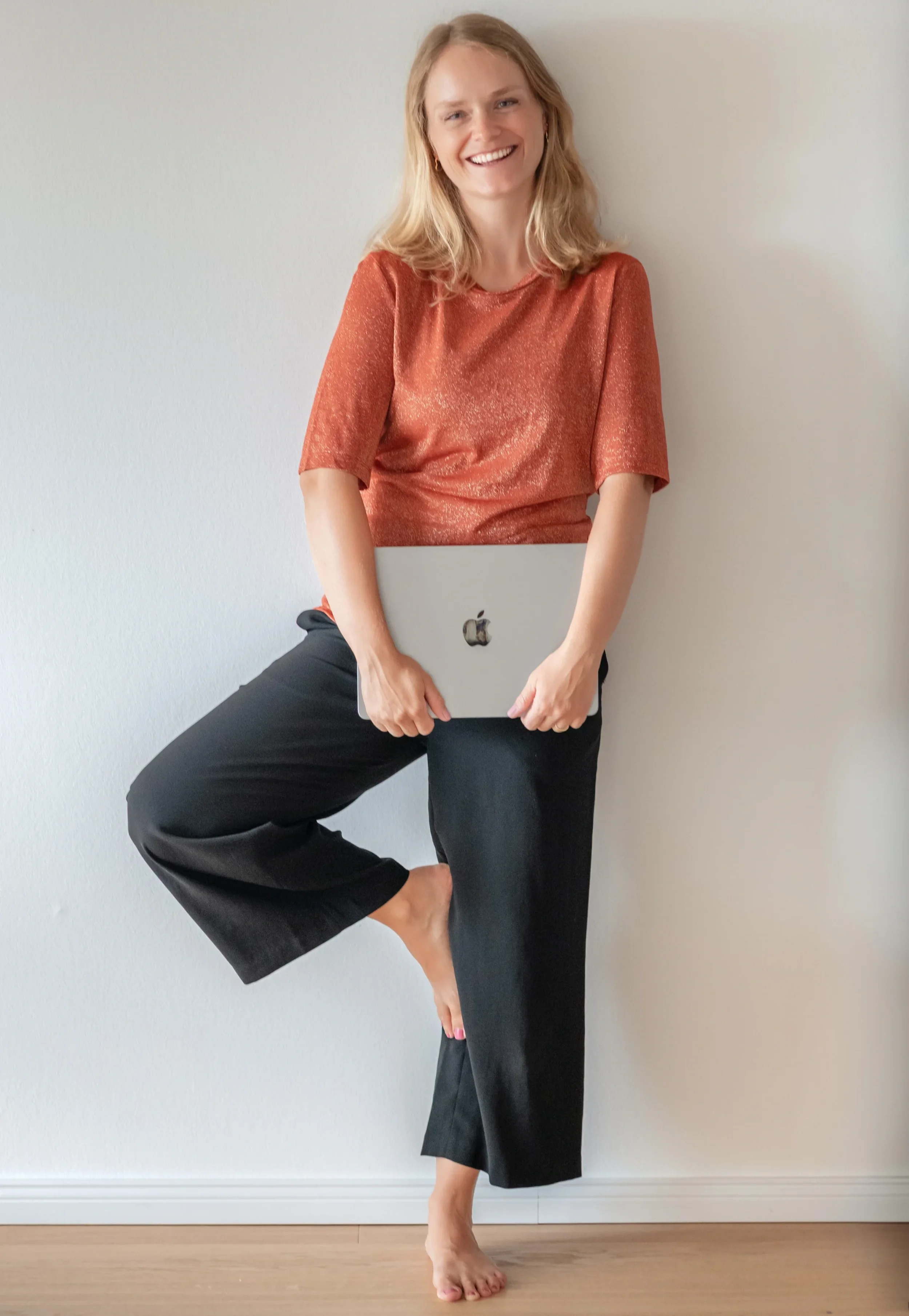 Eine lachende Frau mit lockigem Haar hält ein MacBook in den Händen, steht auf einem Bein, während das andere Bein angewinkelt ist und den Fuß gegen das Knie des stehenden Beins stützt, vor einer weißen Wand.
