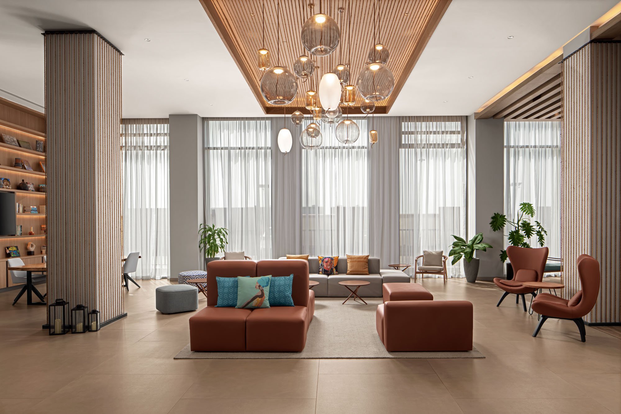 Modern hotel lobby with large windows, sheer curtains, and a variety of seating including armchairs and sofas. Artwork, plants, and a decorative hanging light fixture are visible.