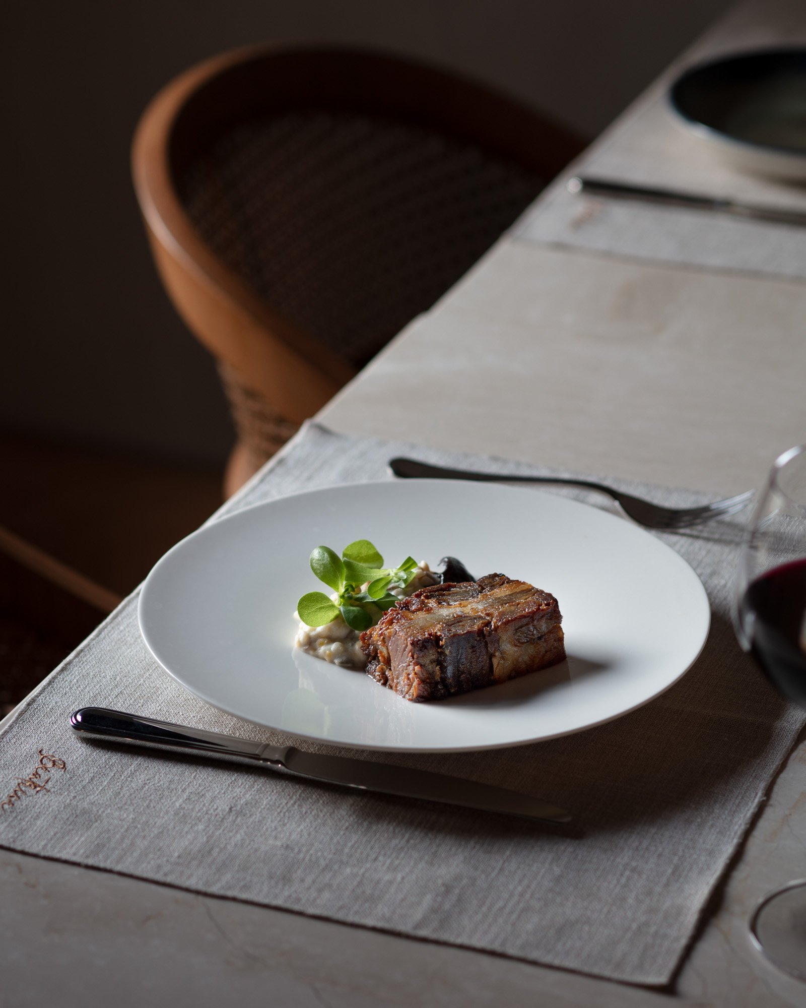 A plated serving of meat with garnish, rice, and sauce, set on a white tablecloth with silverware and a glass of red wine, a chair in the background.