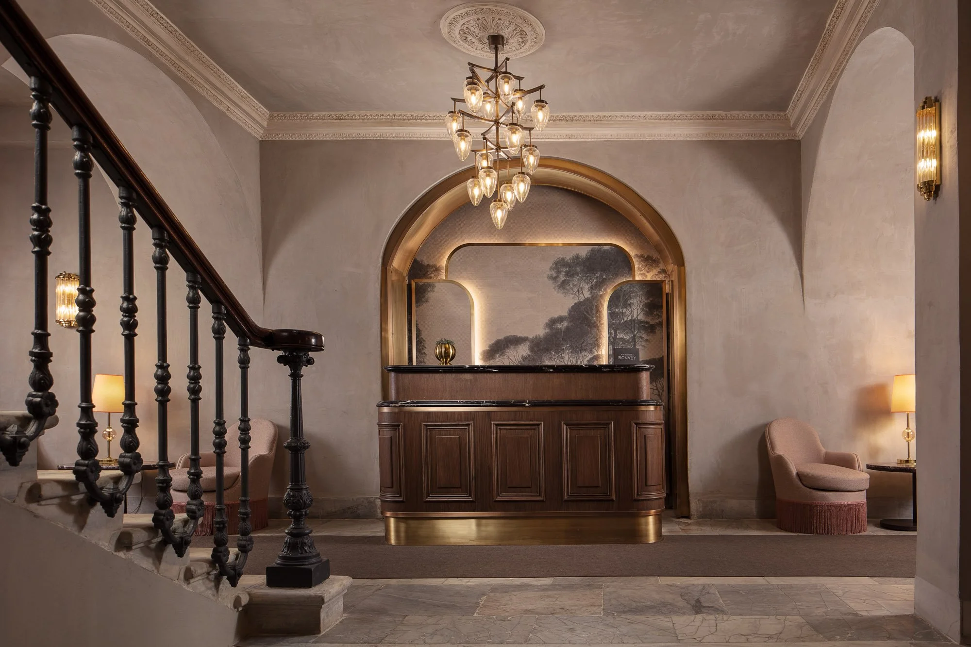 Hotel reception area featuring a brown wooden desk with gold accents, a decorative wall mirror, and a mural of trees behind the desk. Elegant seating includes pink upholstered chairs, with a staircase with ornate black railings on the left and side t