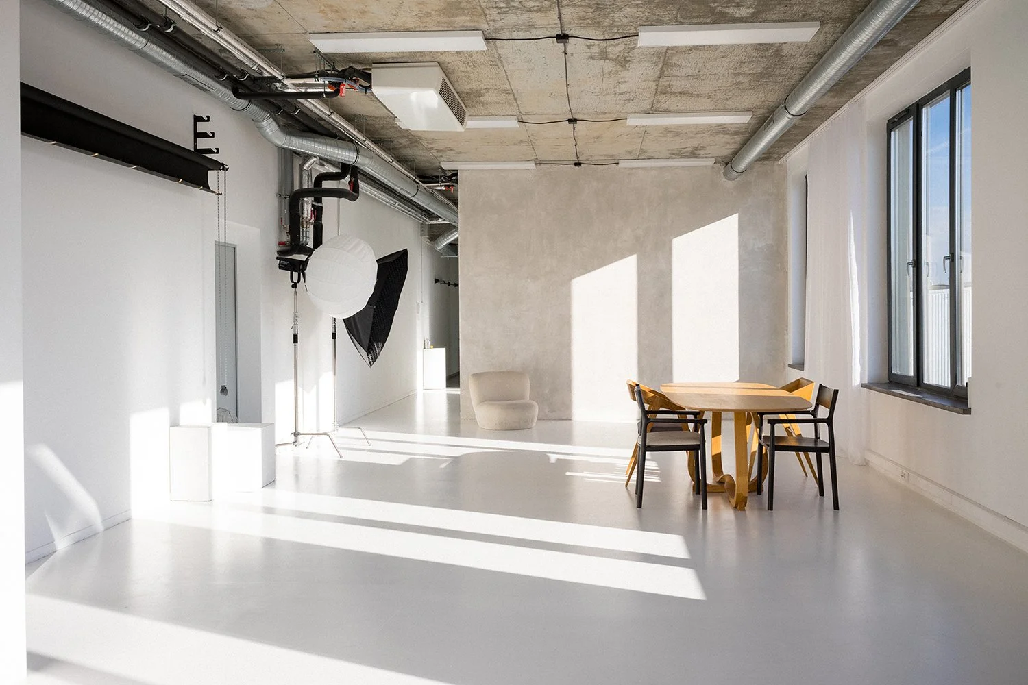 Bright, minimalistic room with white walls and floor, large windows letting in natural light, a wooden dining table with four chairs, a beige armchair, and photo studio equipment including a large softbox and backdrop, in an industrial-style space.