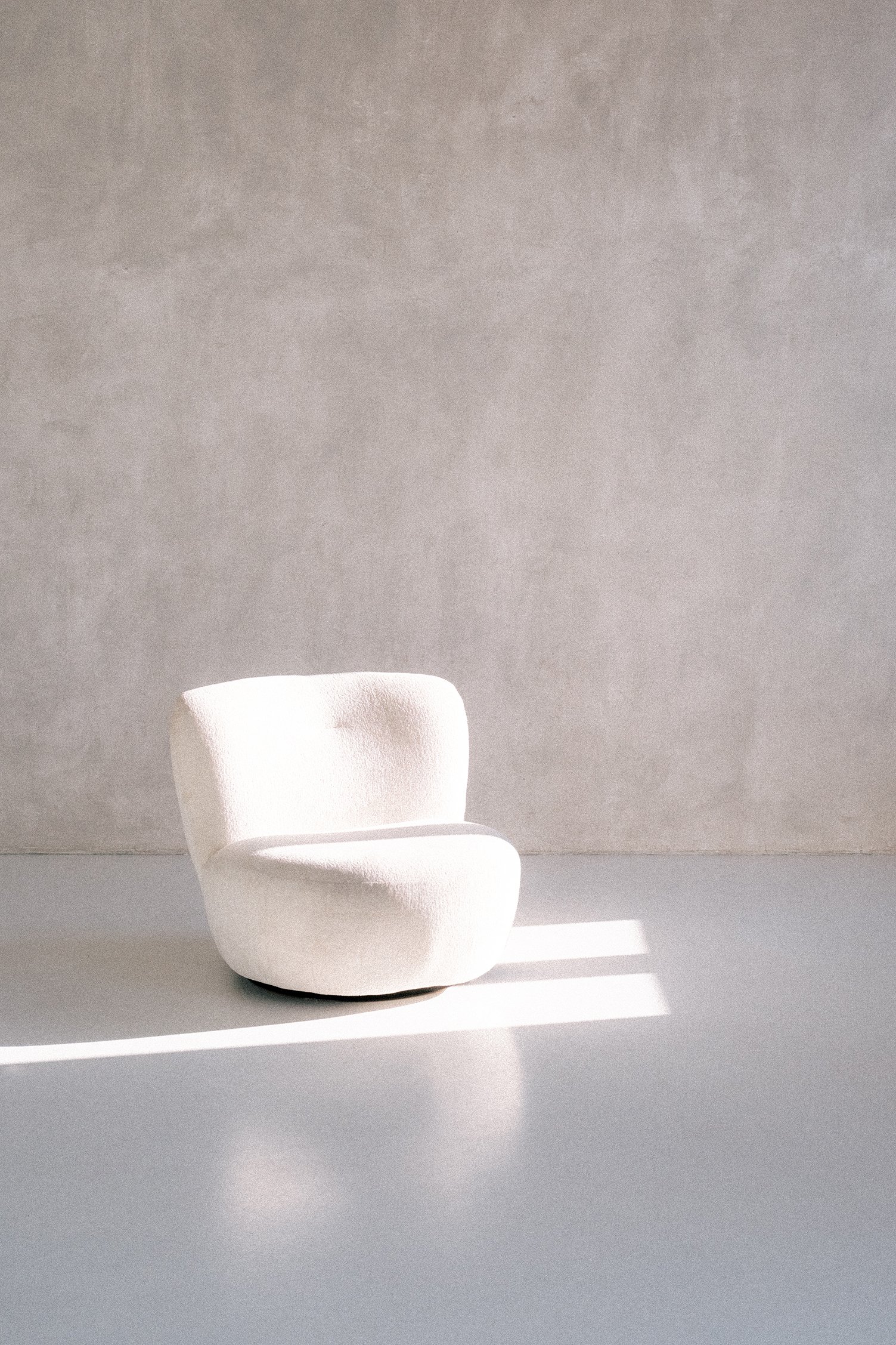 A white, modern, plush armchair in a minimal room with gray walls and sunlight creating diagonal shadows.