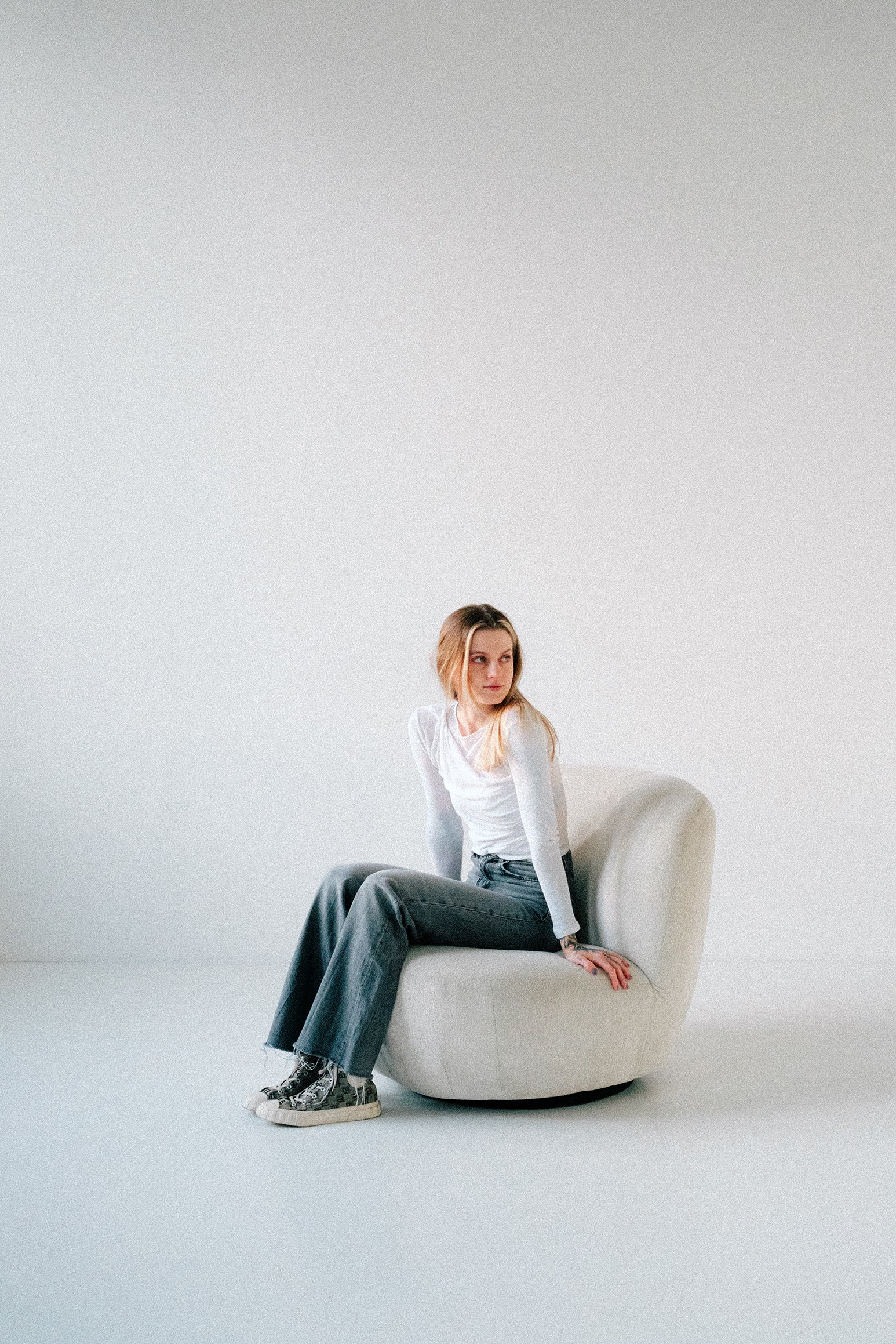 A young woman with blonde hair, wearing a white long-sleeve shirt, dark jeans, and high-top sneakers, sitting on a modern, round, cream-colored chair against a plain white background.