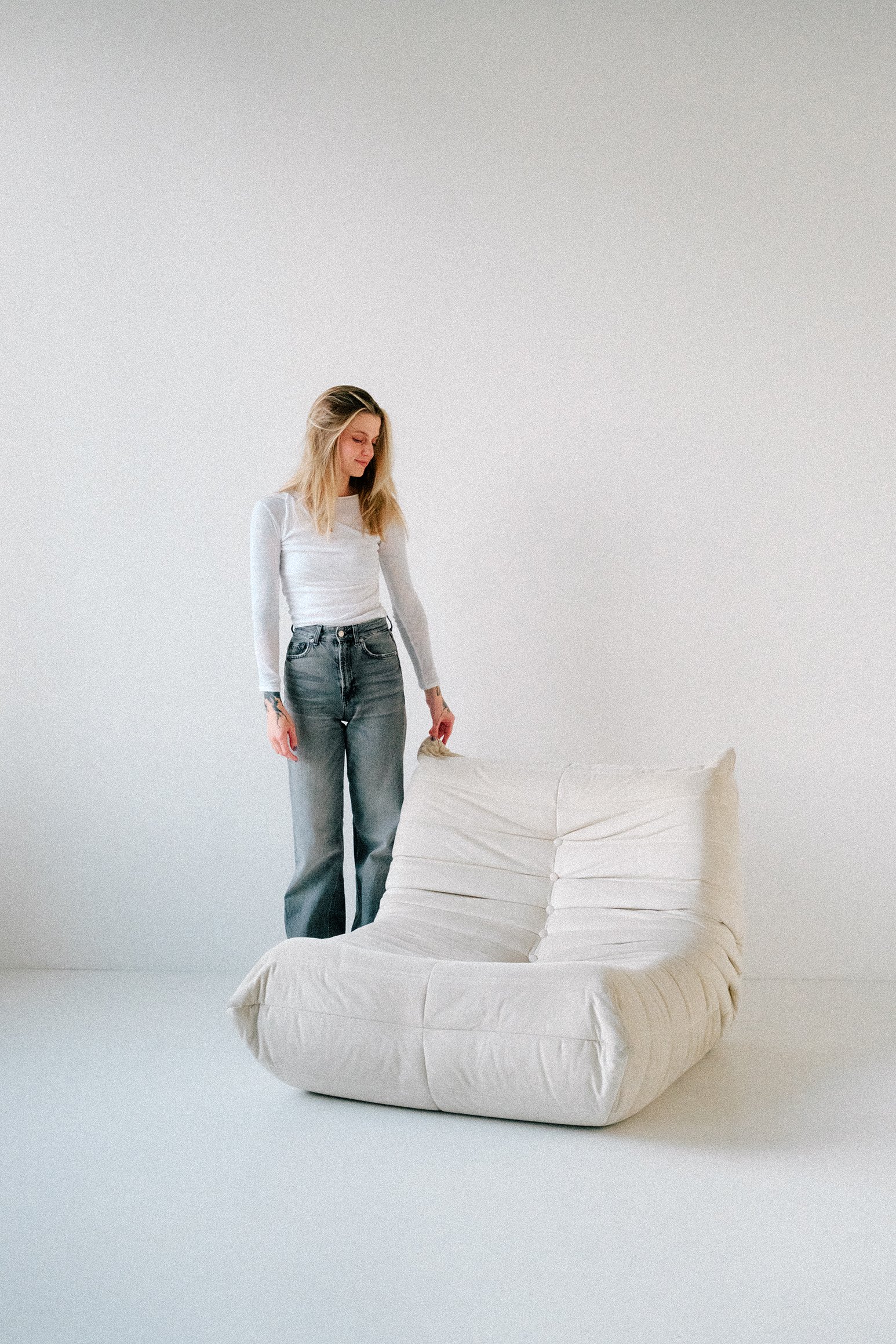 A woman with blonde hair wearing a white long-sleeve shirt and blue jeans standing next to a white, pleated, modern lounge chair in a plain white room.