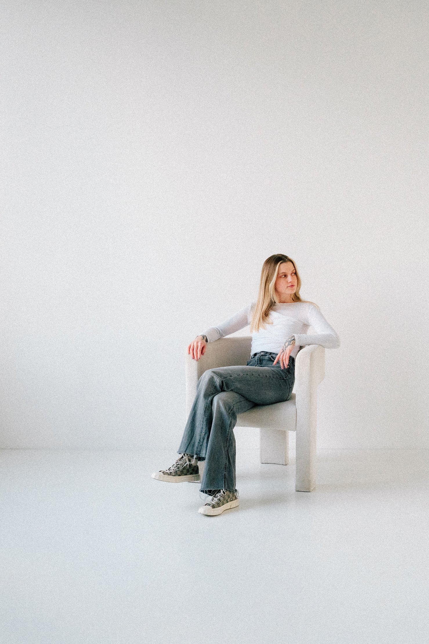 A woman with blonde hair wearing a white long sleeve shirt, gray jeans, and patterned sneakers sitting on a white chair in a minimalist white room.