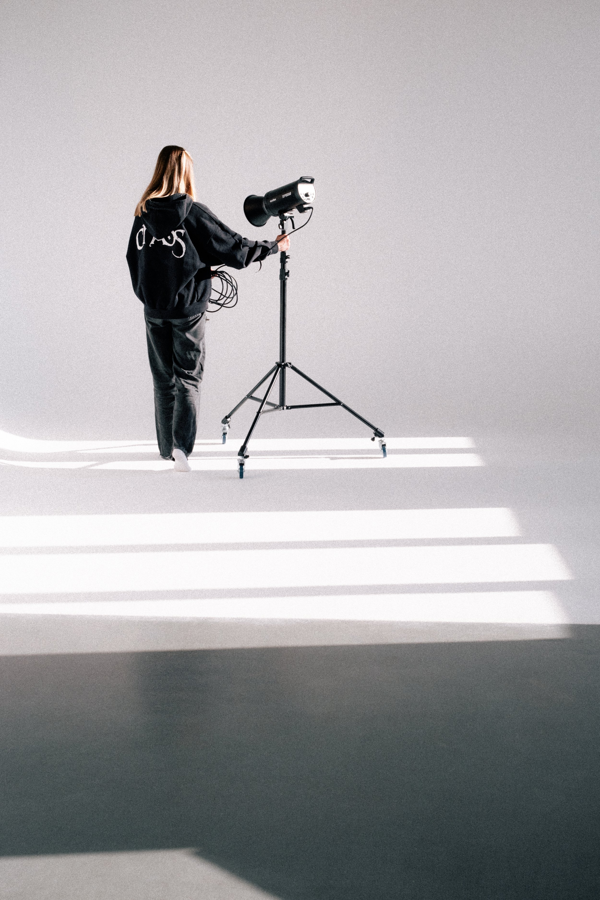 A person wearing a black hoodie with white text on the back adjusting a studio light on a tripod against a plain white background with shadows from window blinds.