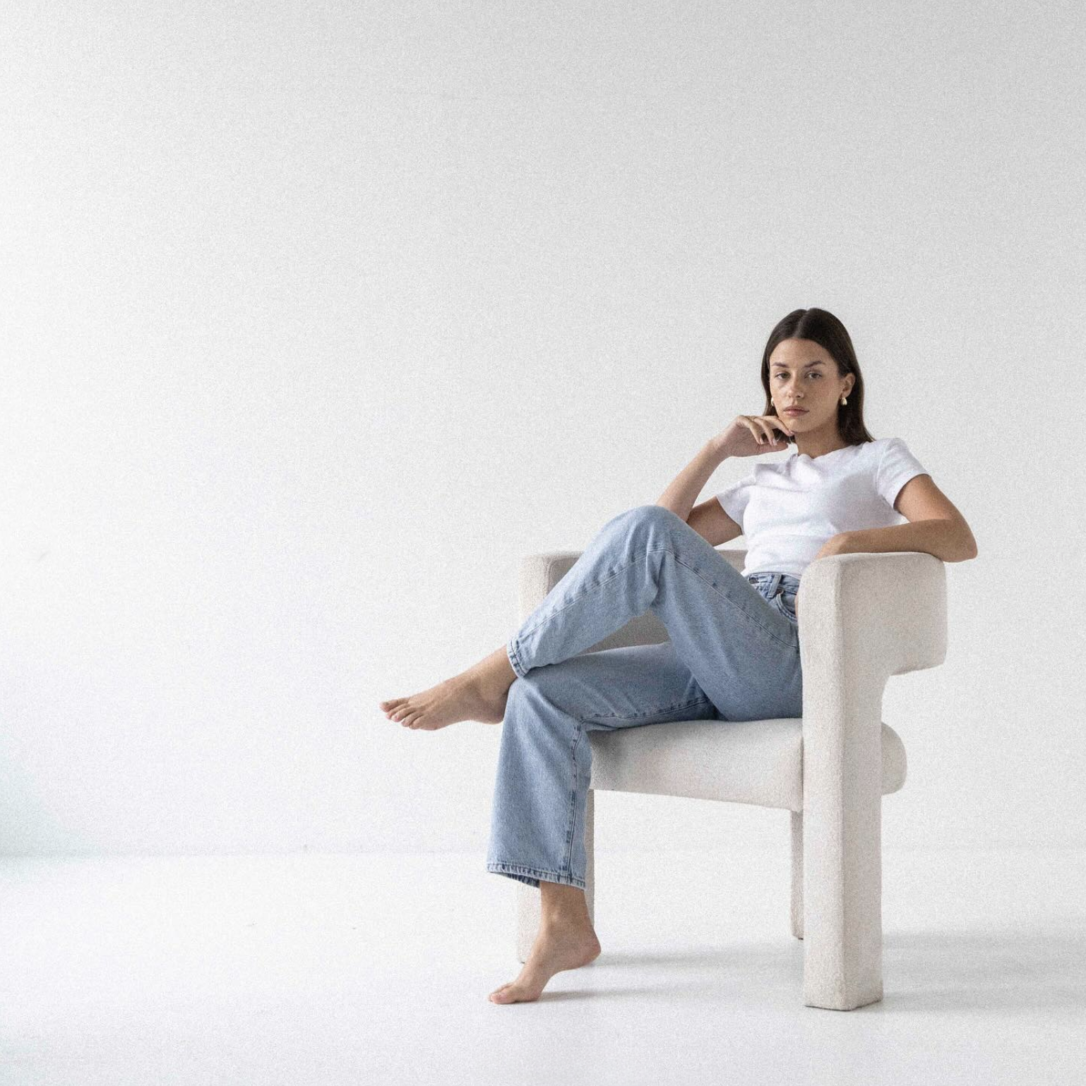 A woman with dark hair sitting in a beige armchair against a plain white background. She is wearing a white t-shirt and light blue jeans, with one leg crossed over the other and her right hand resting near her chin.