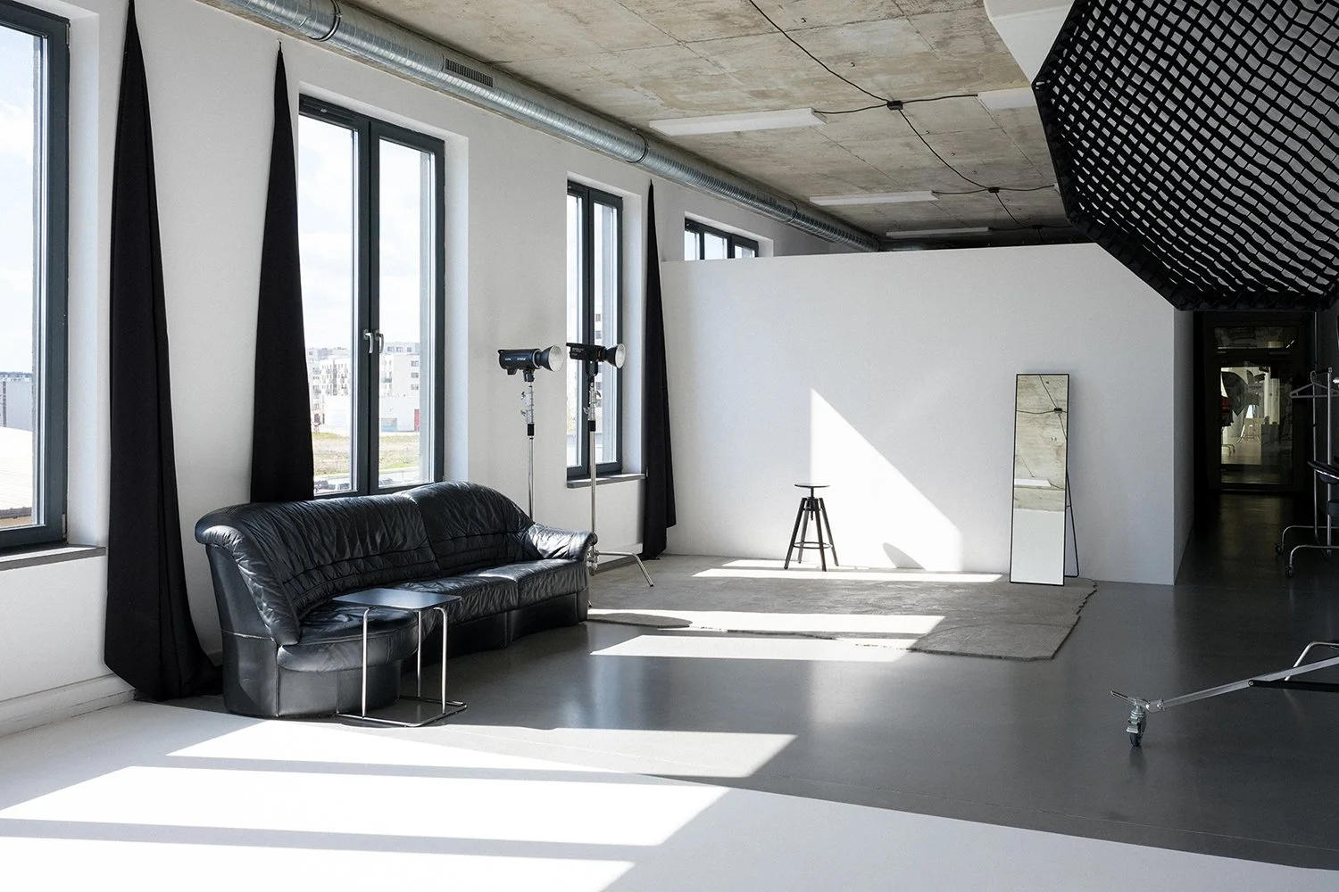 A modern photography studio with large windows, black curtains, a black leather sofa, light stands, a full-length mirror, a rug, and a backdrop wall. The ceiling has an industrial look with exposed concrete and ventilation pipes.