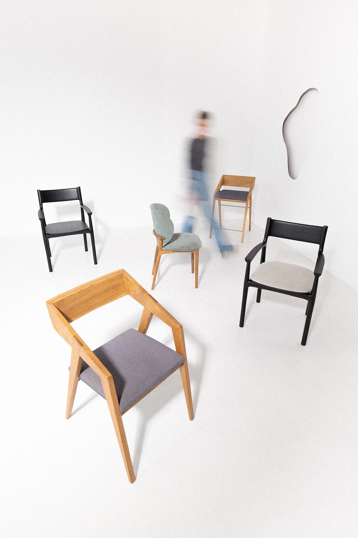 Multiple modern chairs of various designs and colors are arranged on a white floor with a plain white wall background. A blurred person walks among the chairs.