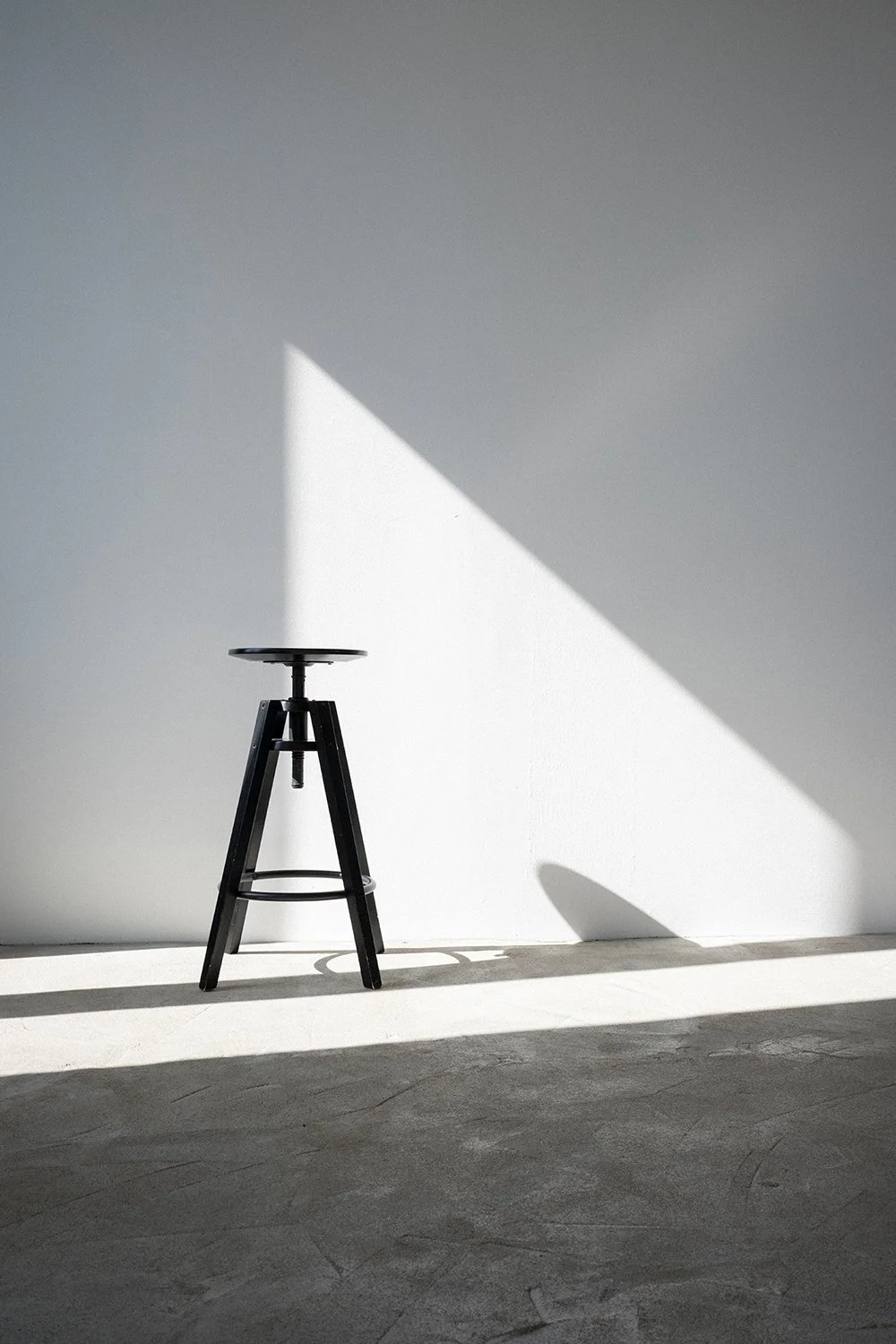 A black adjustable stool positioned against a plain white wall, with bright sunlight casting a shadow on the wall and floor.