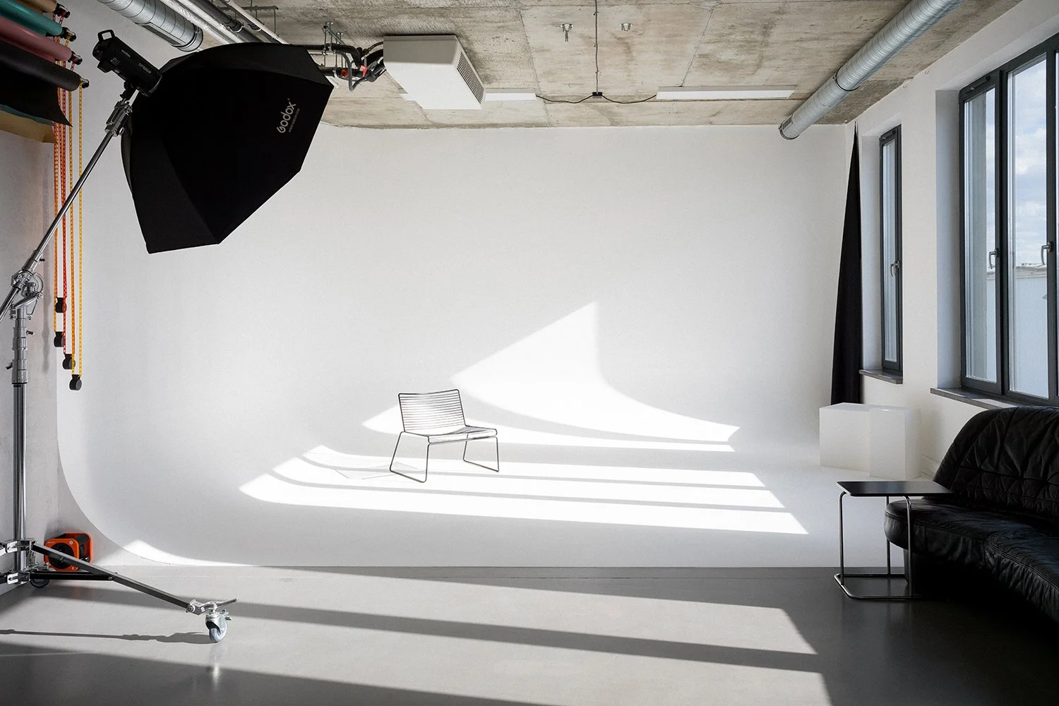 A photography studio with a white backdrop, a black leather couch, a small table, a black seating chair, and large windows allowing natural light. Photography equipment, including a light with a softbox and a stand, is visible.