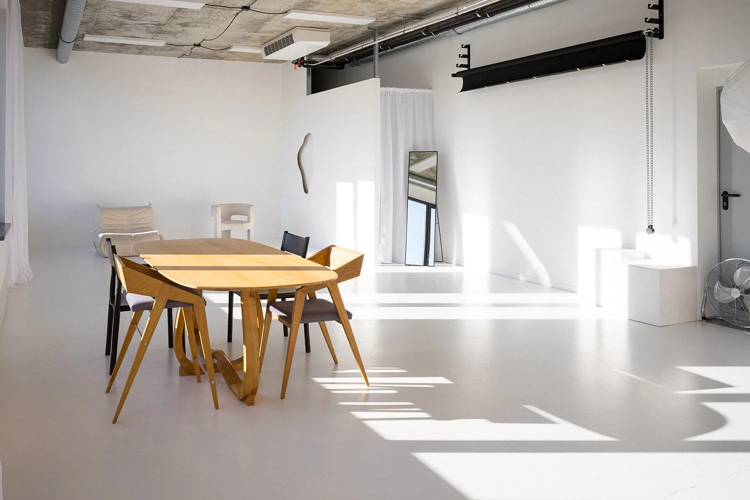 Interior of a minimalistic, modern room with white walls and floor, including a wooden dining table with six chairs, a large mirror leaning against the wall, a white curtain, and a wall decoration. Sunlight streams through windows casting shadows on 