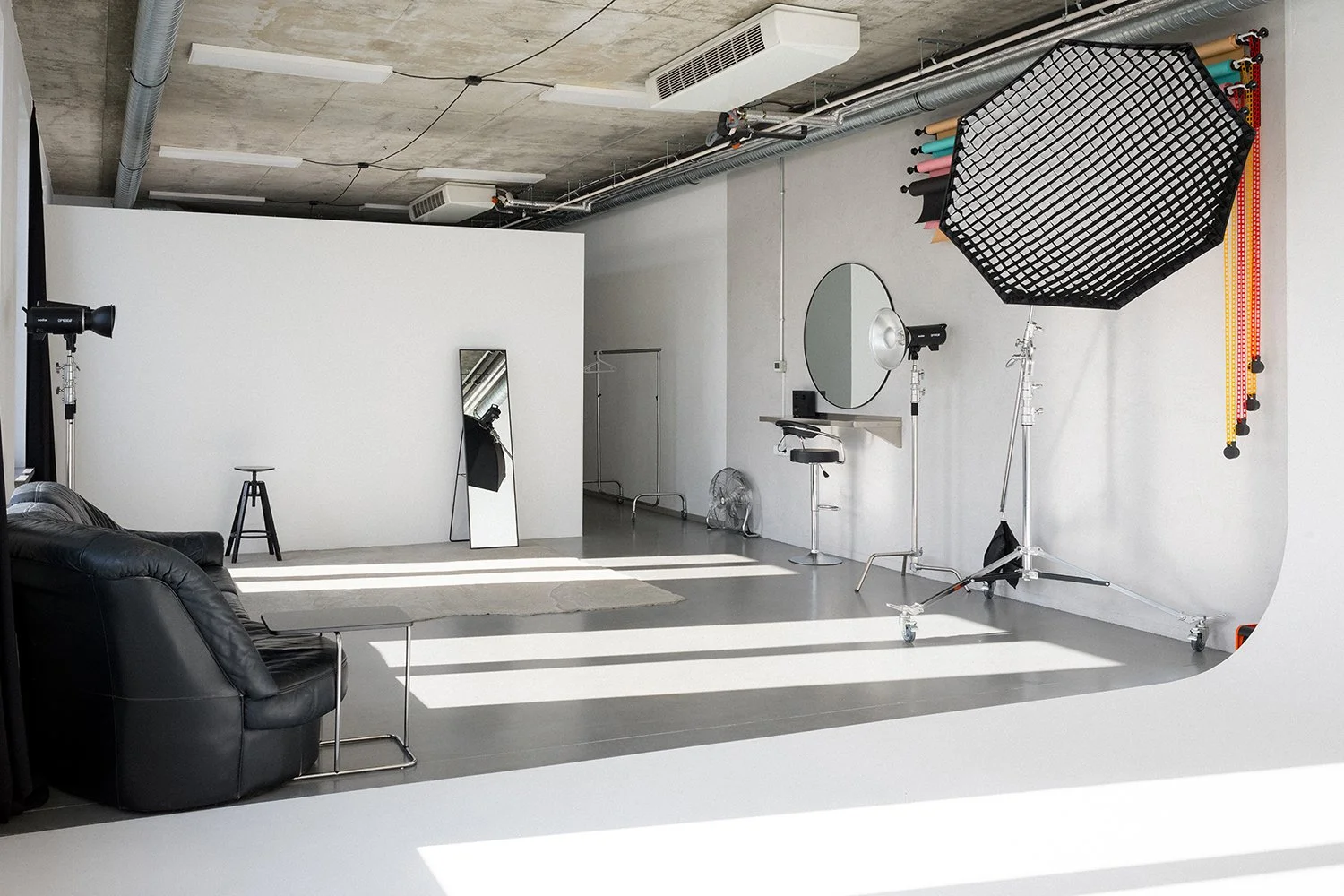Photo of a photography studio with a black leather sofa, a small side table, and a full-length mirror leaning against a white wall. The studio has professional lighting equipment, a large umbrella reflector, and various lighting stands. There is a wi