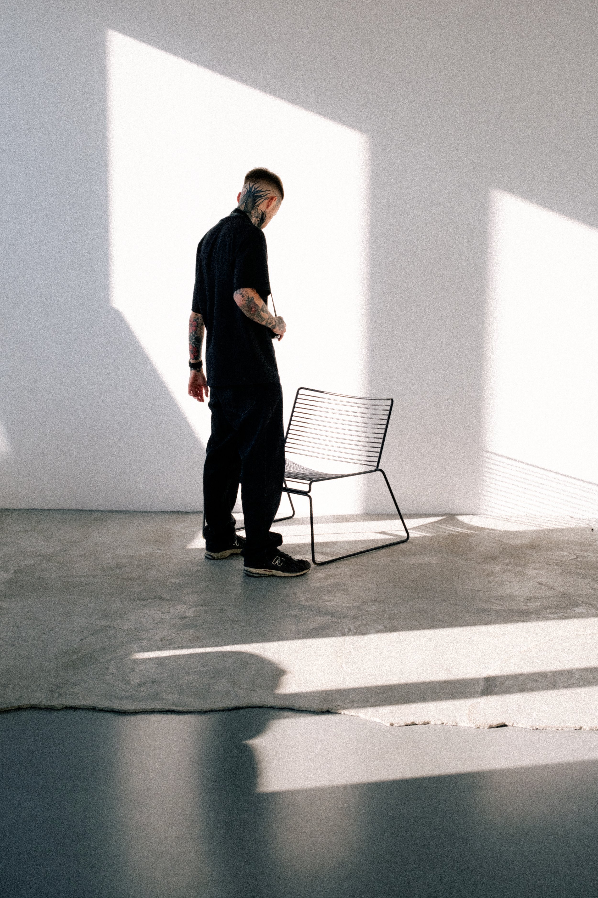 Man with tattoos standing in a minimalistic room with sunlight casting shadows, next to a black wire chair.