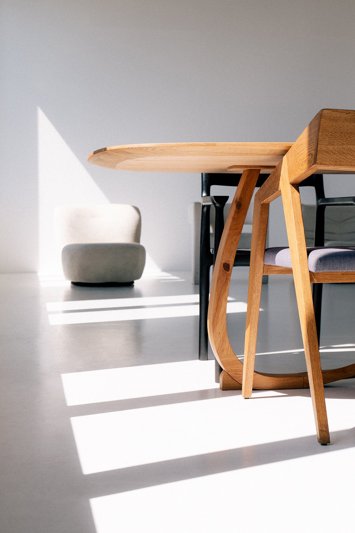 A minimalist dining room with a wooden table, gray chair, and sunlight casting shadows on the white floor next to a white and gray armchair in the background.