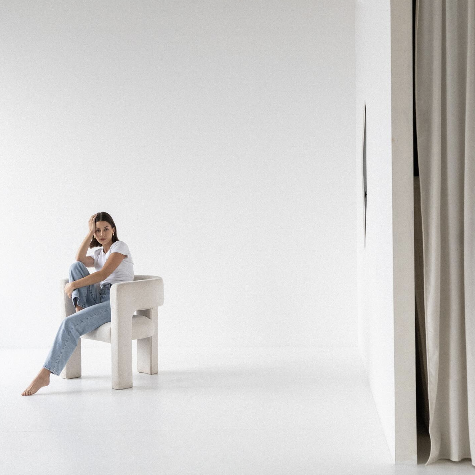 A woman sitting on a white chair in a minimalistic, all-white room, with her right arm resting on her knee and her left hand touching her head, wearing a white t-shirt and jeans.