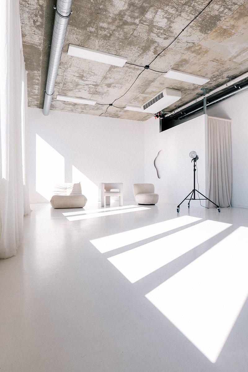 Bright photography studio with white walls, minimal furniture, and sunlight casting shadows through curtains onto the floor. There is a studio light on a stand, a chair, a sofa, industrial piping on the ceiling, and a ceiling vent.