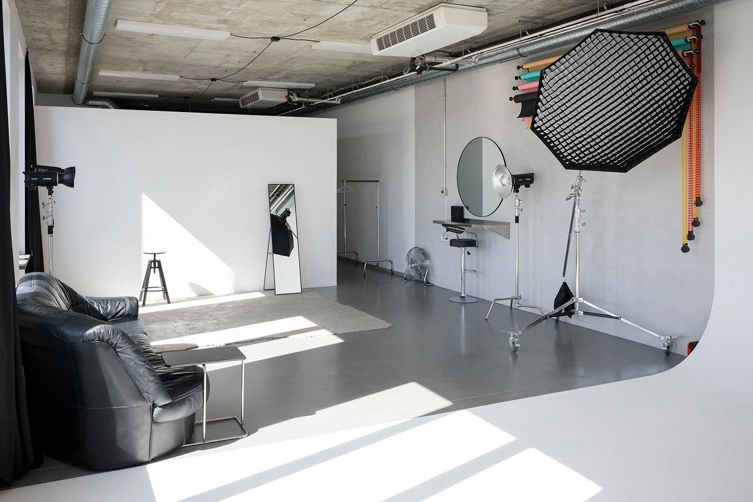 Professional photography studio with a black leather couch, a small table, a stool, a mirror, lighting equipment, and backdrops.
