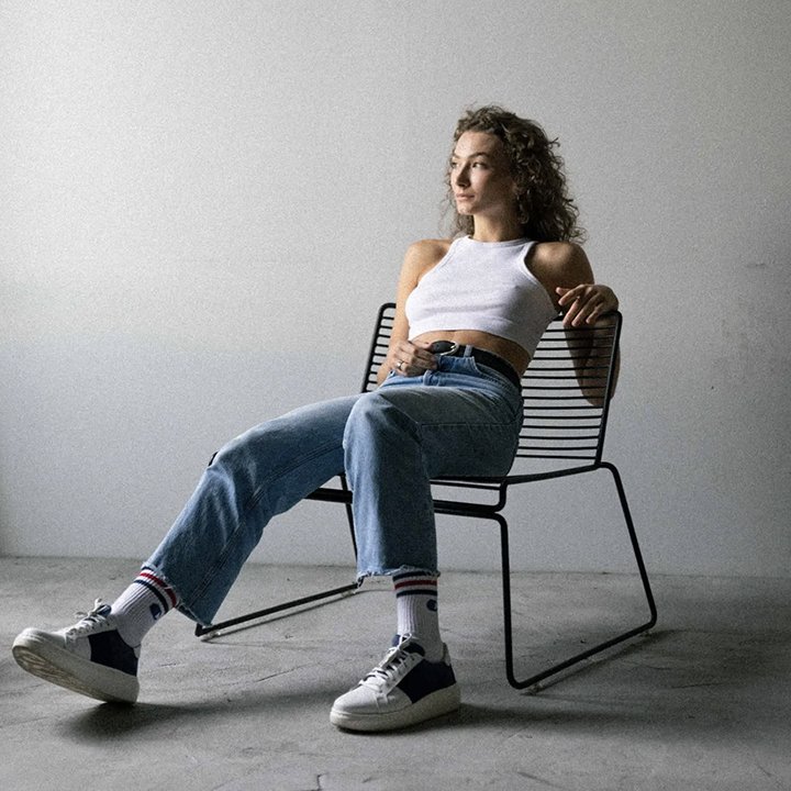 Young woman with curly hair sitting on a modern black metal chair against a plain white wall, wearing a white tank top, blue jeans, and sneakers with striped socks.