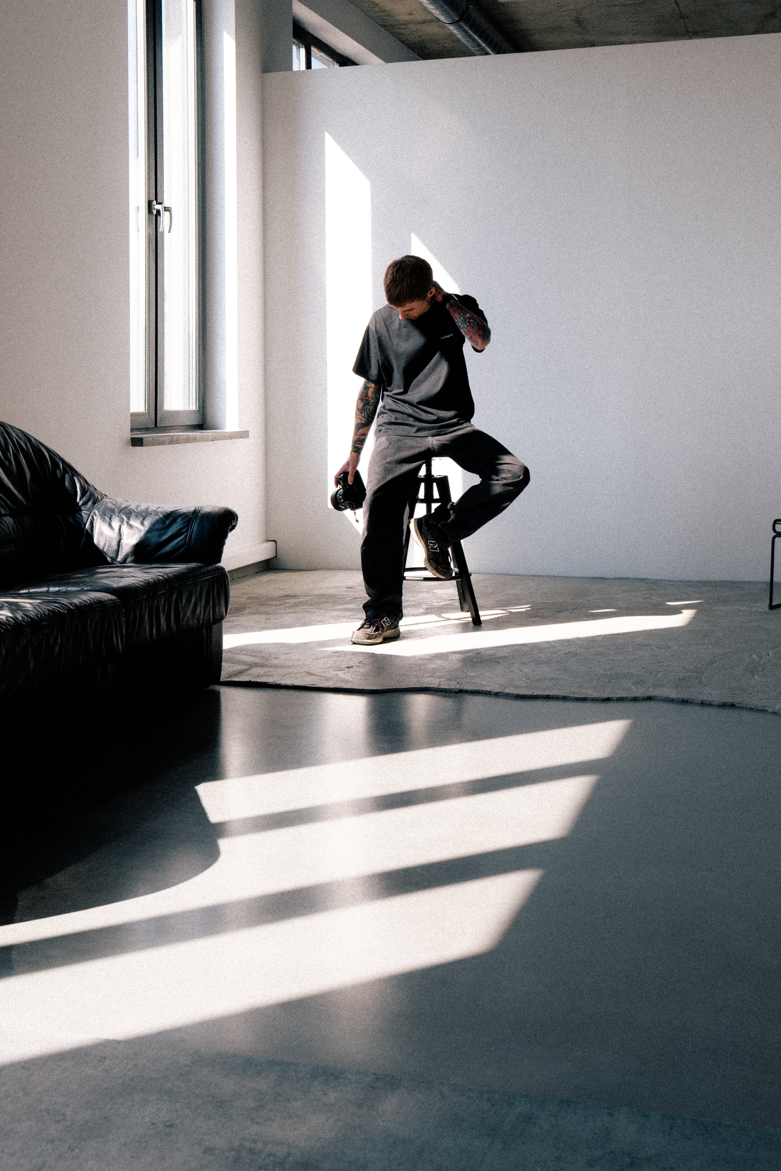 A young man with tattoos, wearing a black t-shirt and dark pants, sitting on a stool in a bright, minimalist room with sunlight streaming in through a large window, holding a camera in his hand.