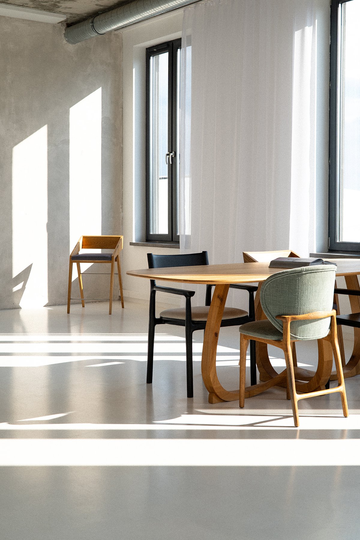 Minimalist dining area with a wooden table, four chairs, large windows, sheer curtains, and sunlight casting shadows on the white floor.