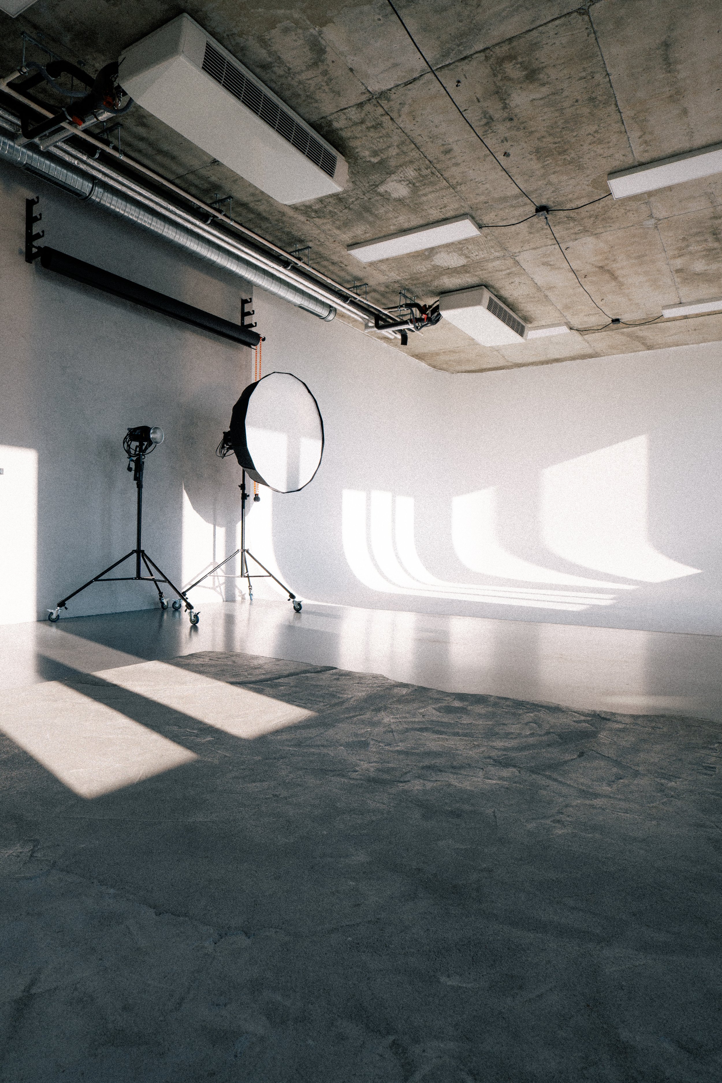 Photo of a photography studio with lighting equipment, a white backdrop, and shadows cast on the wall by sunlight.
