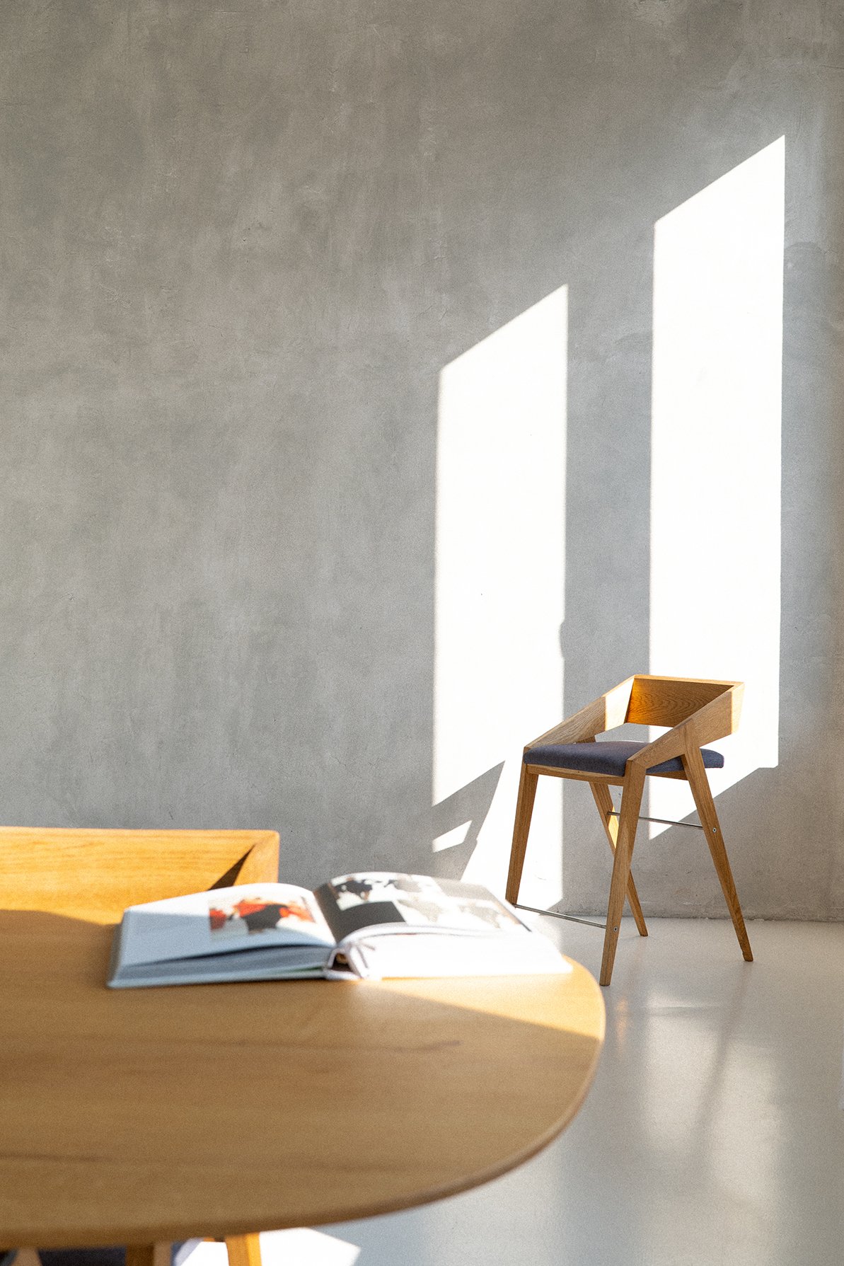 Bright sunlight casting geometric shadows through a window onto a minimalist room with a wooden chair and an open magazine on a wooden table.