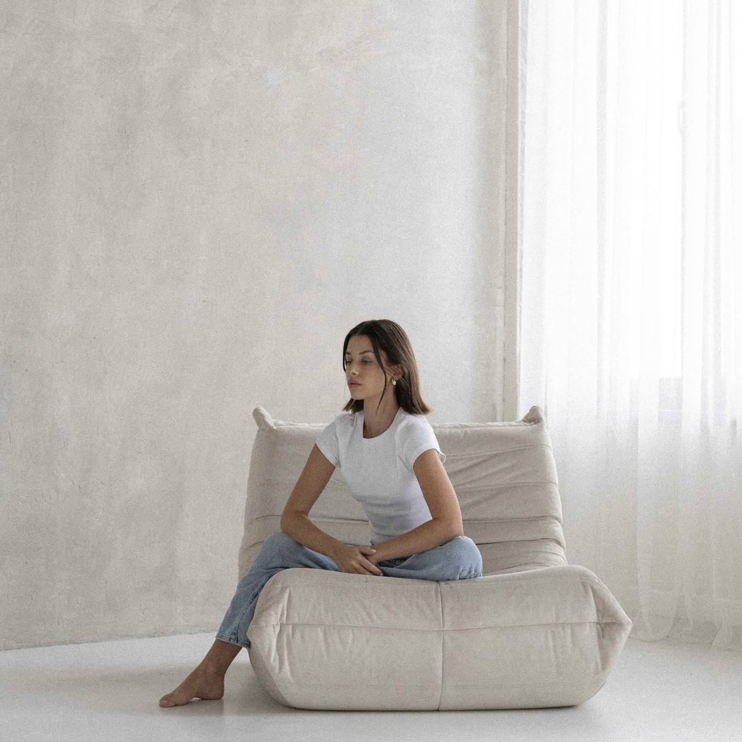 A woman sitting cross-legged on a beige sofa in a bright, minimalist room, wearing a white t-shirt and light blue jeans, with her eyes closed and hands resting on her lap.