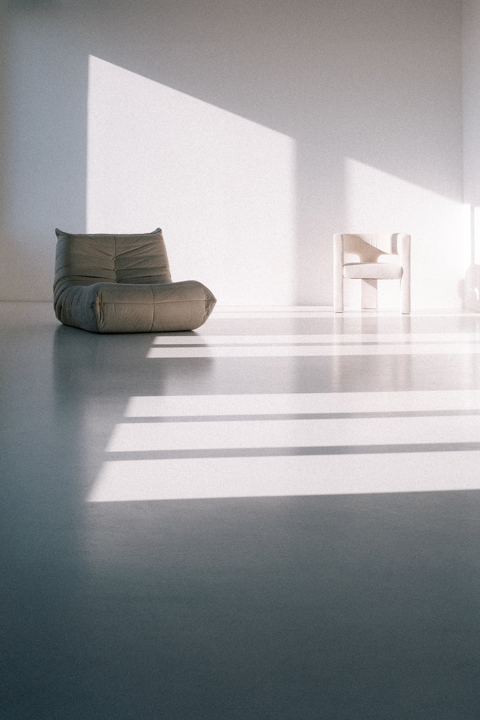 Minimalist room with a brown cushion chair and a white armchair, both casting shadows on a white floor due to sunlight through blinds.