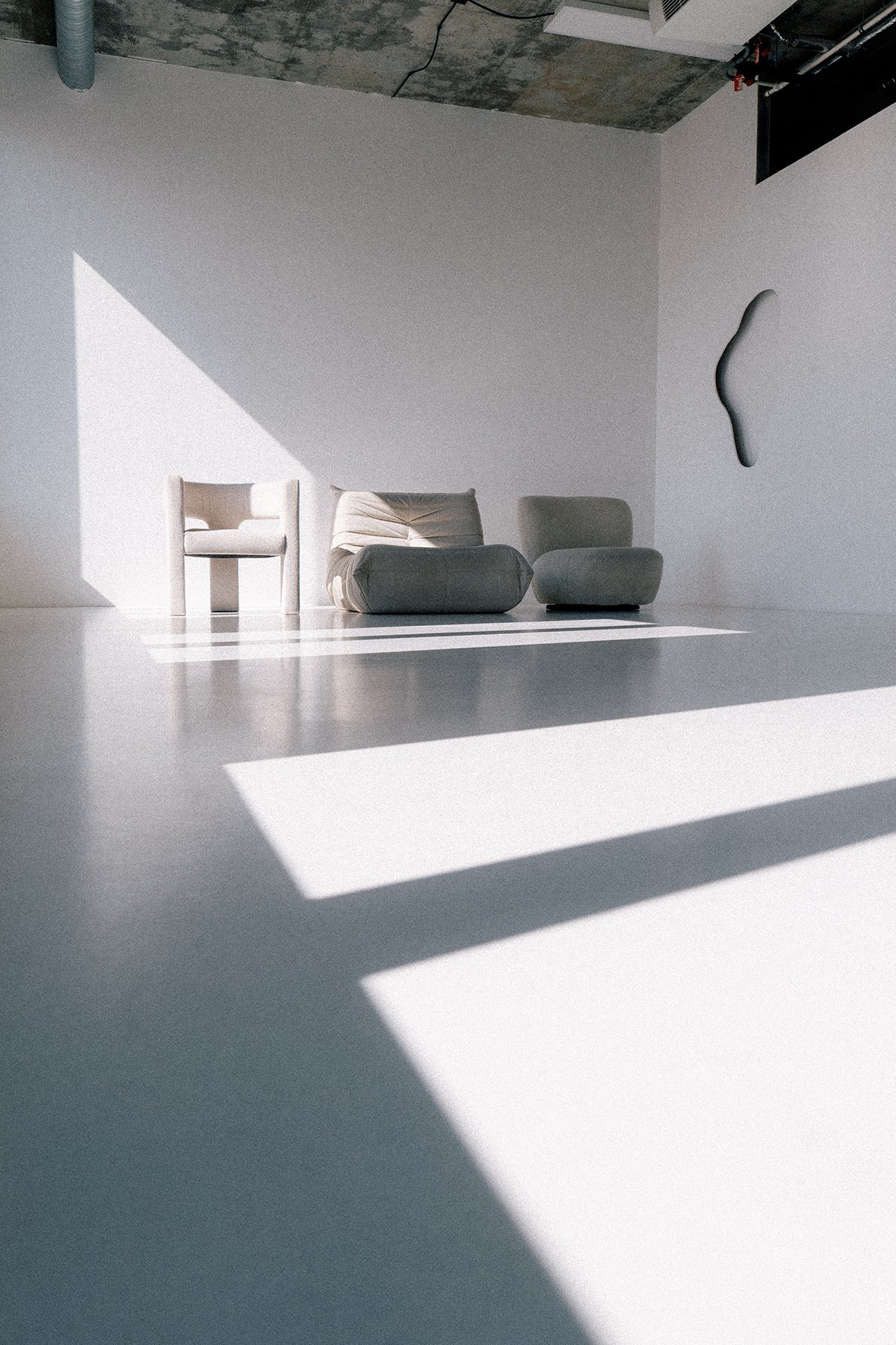 Minimalist interior with three modern chairs and wall sculpture, sunlight creating geometric shadow patterns on light-colored floor and wall.