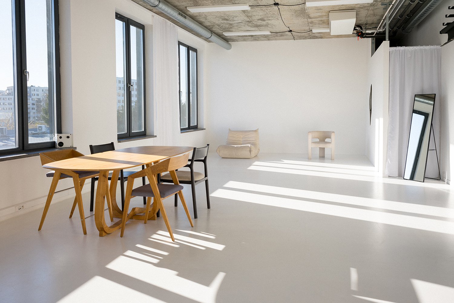 Minimalist, modern room with white walls and floor, three tall windows letting in natural light, a wooden dining table with four chairs, a beige beanbag, a white chair, a tall mirror leaning against the wall, and a large mirror with a white curtain h