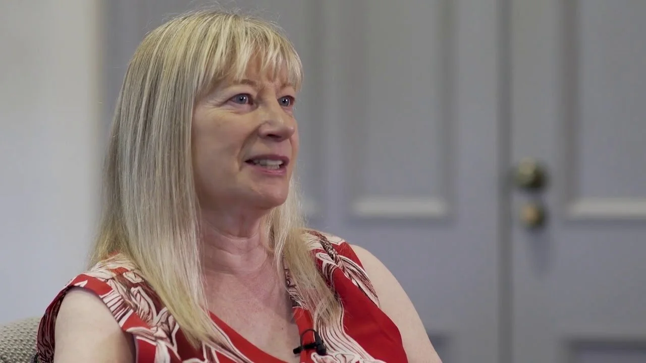 A middle-aged woman with blonde hair wearing a red and white patterned sleeveless top, speaking in conversation.