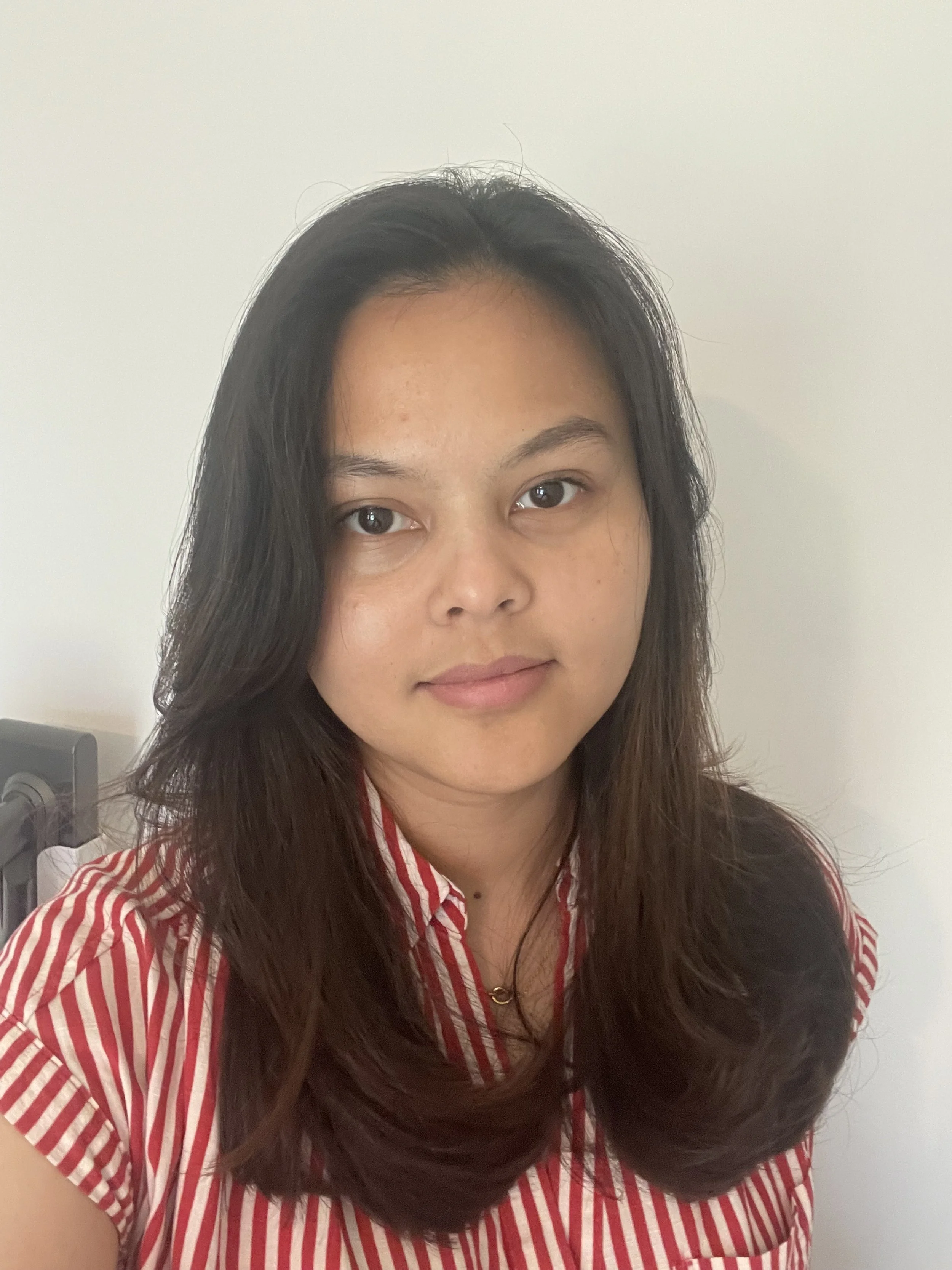 A woman with long dark hair wearing a red and white striped shirt, sitting against a plain white wall.