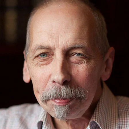 Close-up of an older man with a grey beard and moustache, wearing a checkered shirt, looking at the camera.
