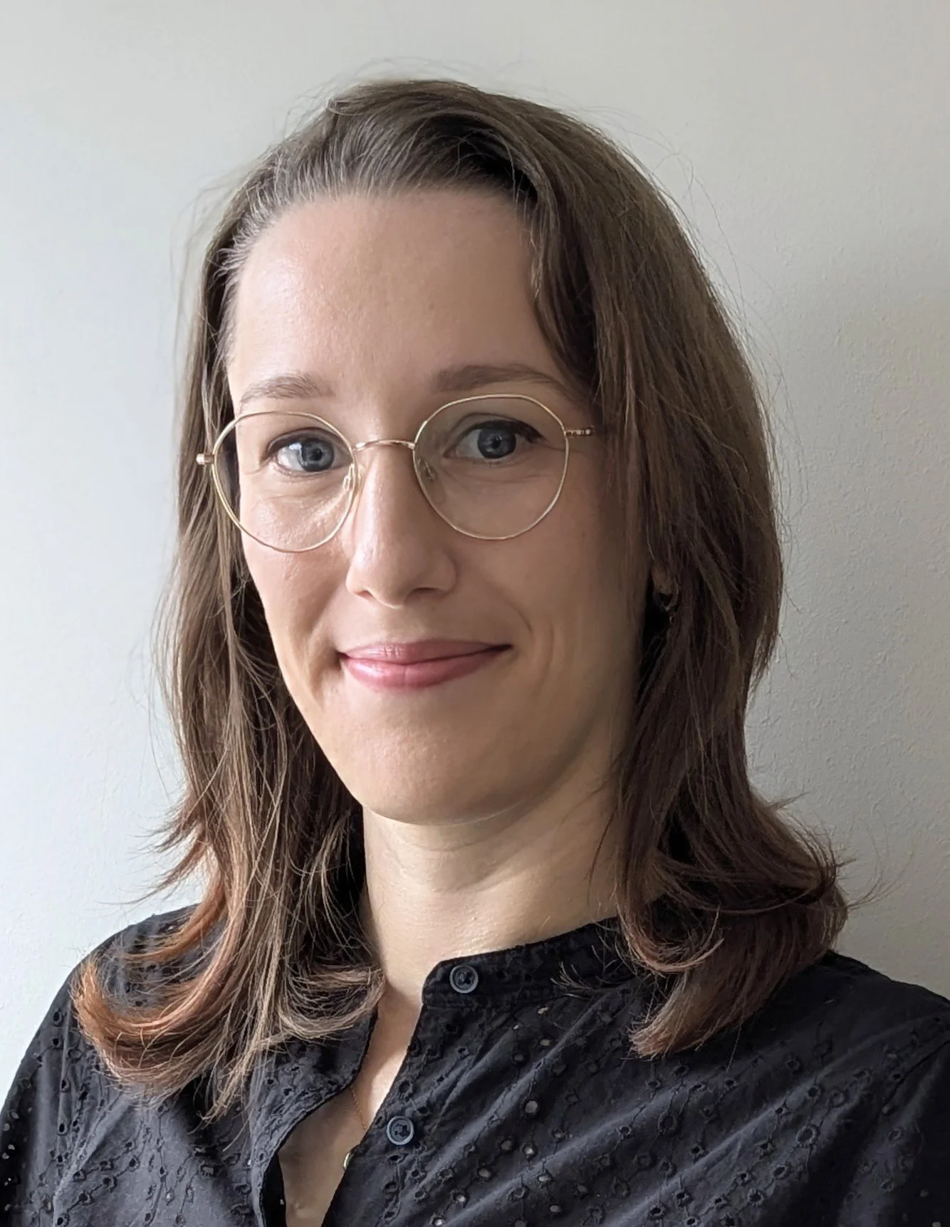 A woman with shoulder-length brown hair, blue eyes, wearing glasses and a black patterned top, smiling in front of a plain white wall.