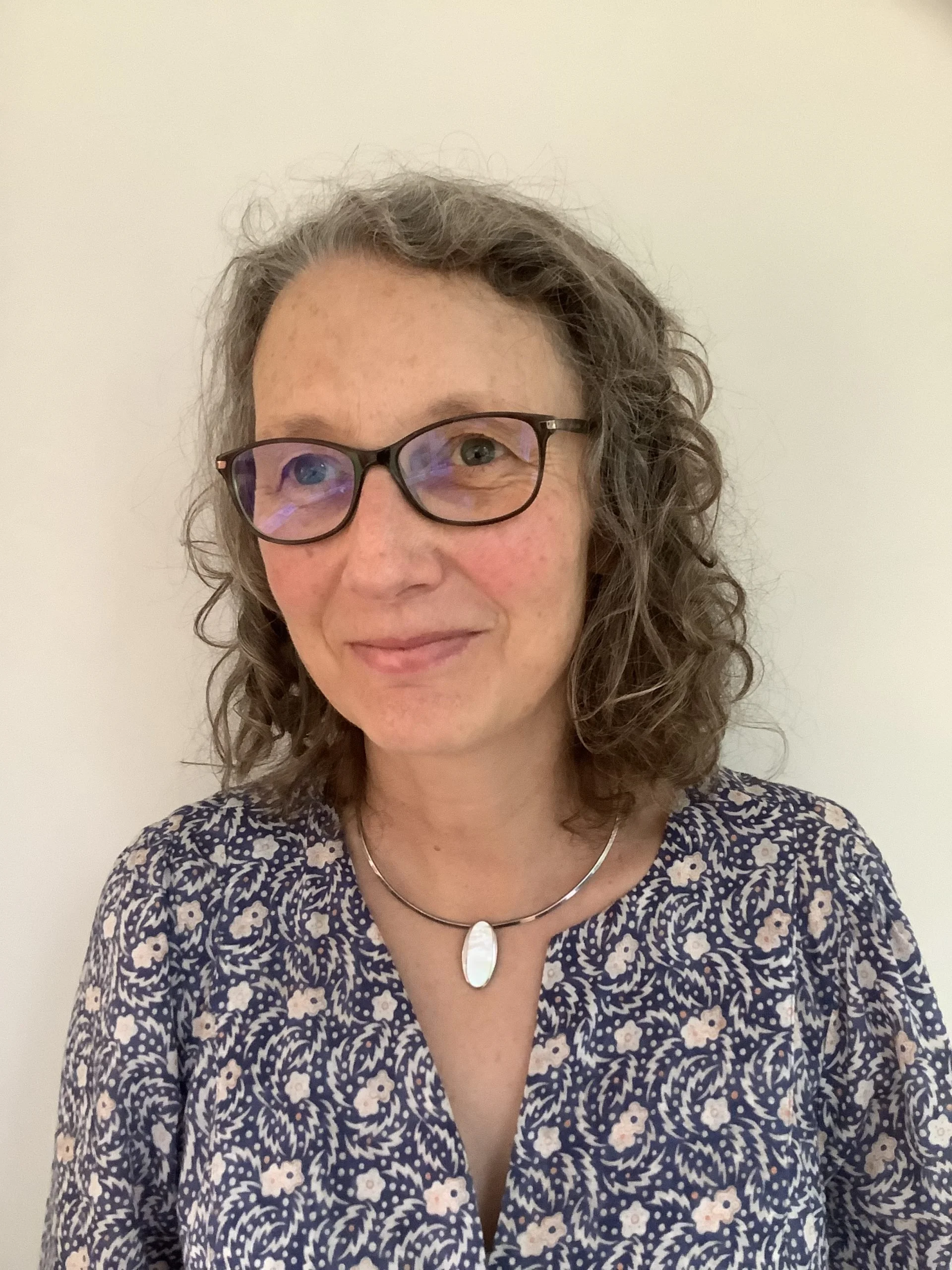 A woman with shoulder-length curly hair, glasses, and a necklace, wearing a floral-patterned blouse, standing against a plain white wall.