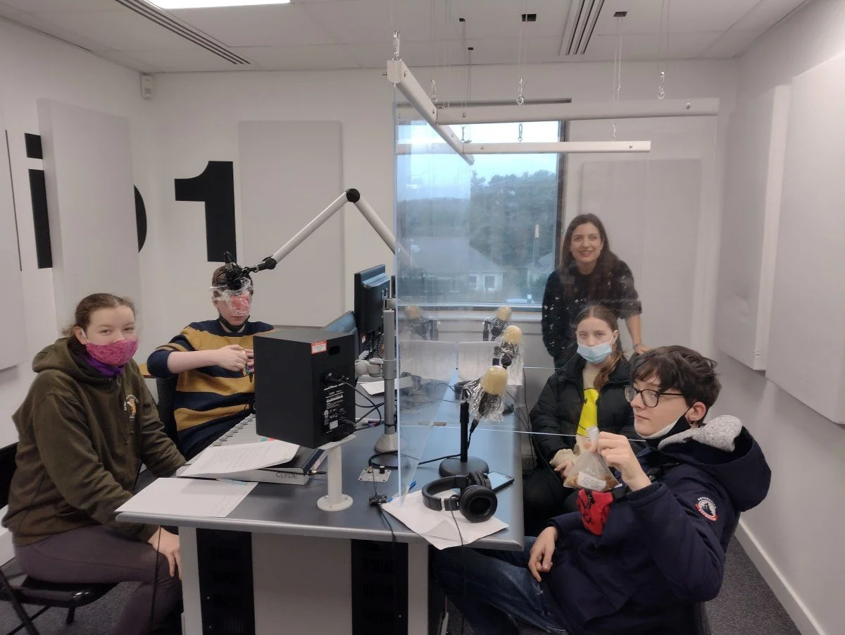Group of five people, four young students and one adult, sitting in a recording studio with microphones, a computer, and soundproofing walls, some wearing masks and one person holding a snack.