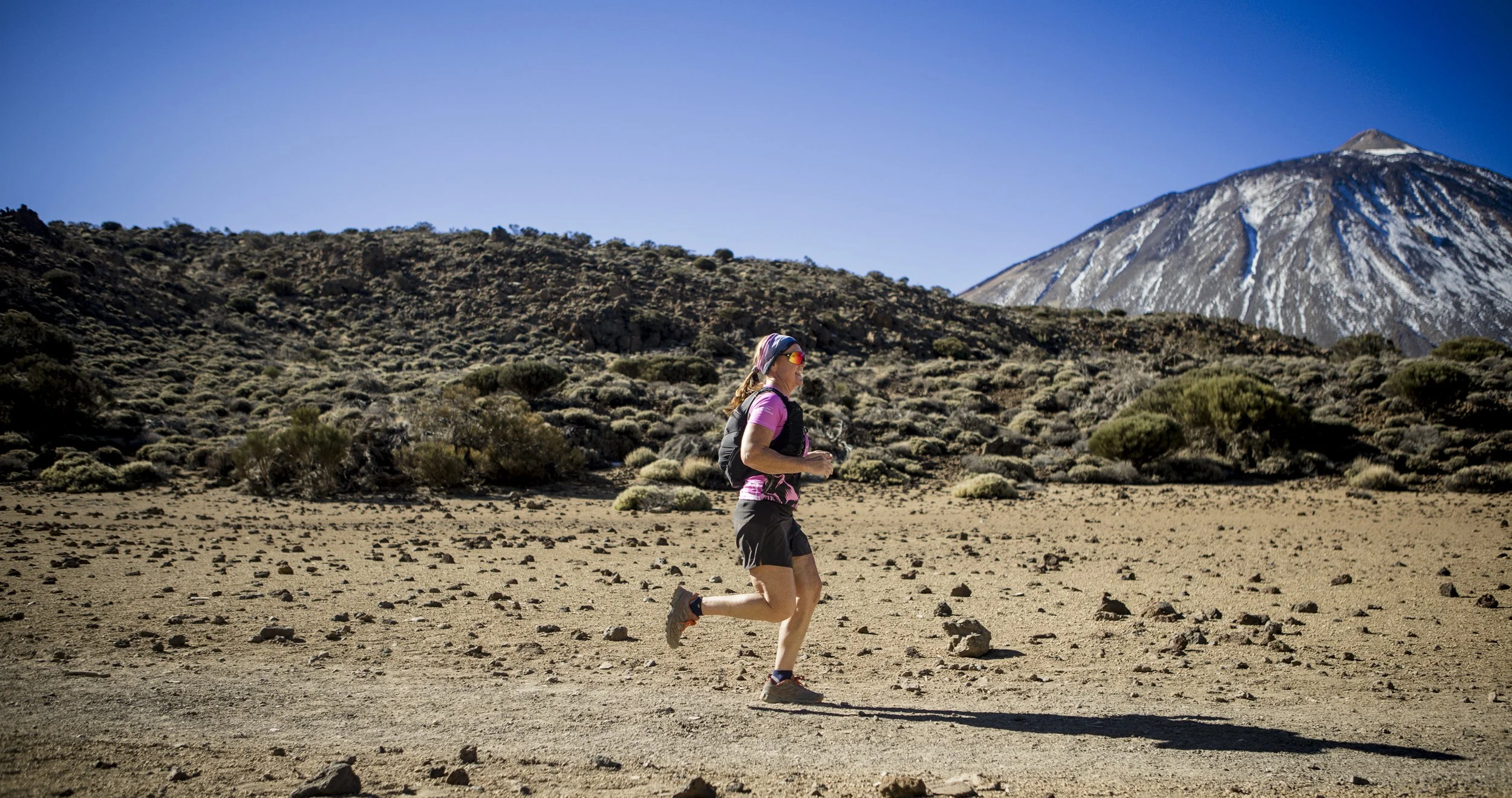 Leanne Rive trail running on a dirt path in a mountainous, desert landscape with sparse bushes and a snow-capped volcano in the background under a clear blue sky.