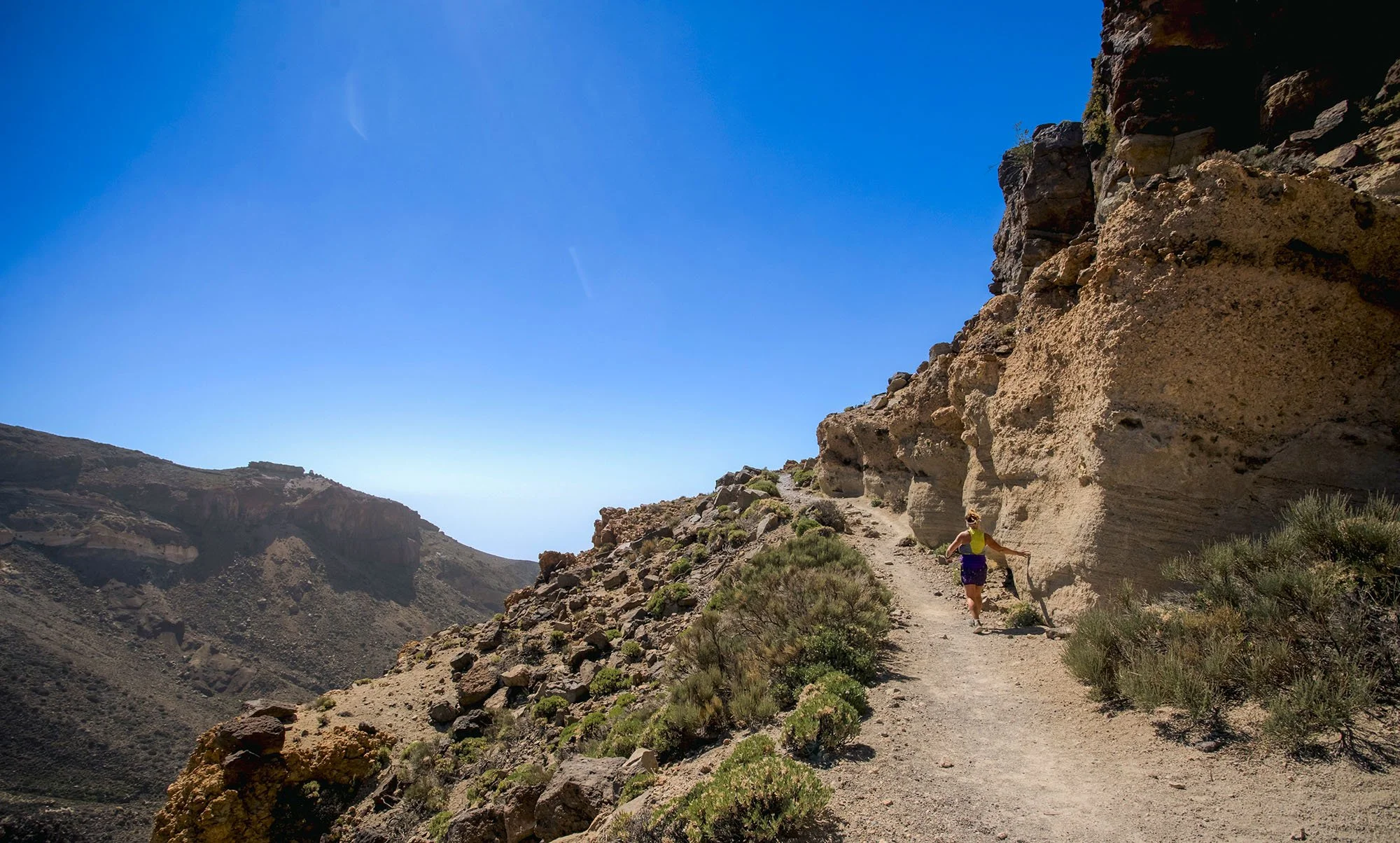 A person hiking along a rocky mountain trail with a steep cliff on one side, under a clear blue sky.