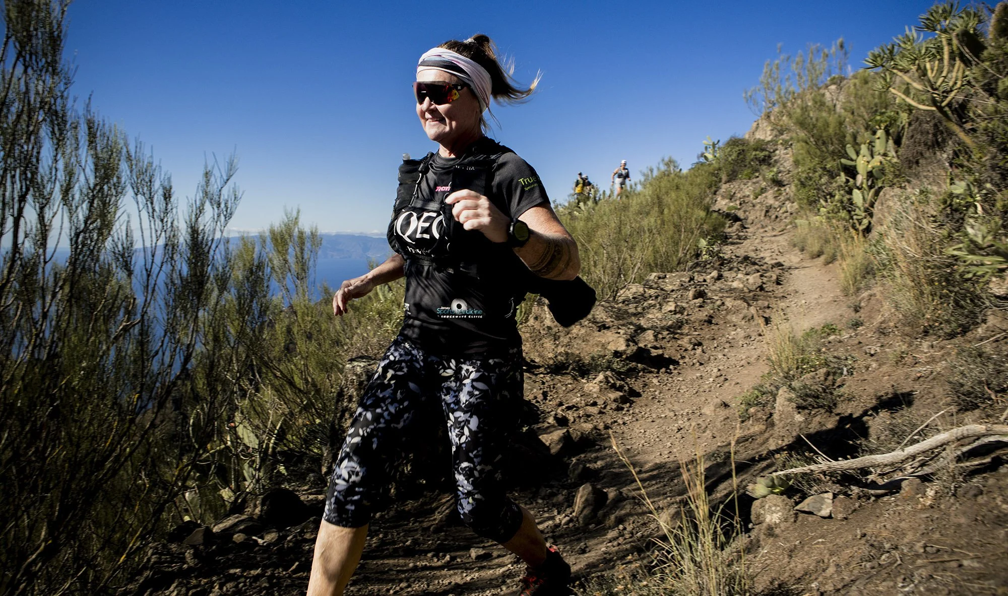 Leanne Rive trail running on a dirt mountain path with a scenic view of blue sky in Tenerife, mountains, and vegetation in the background.