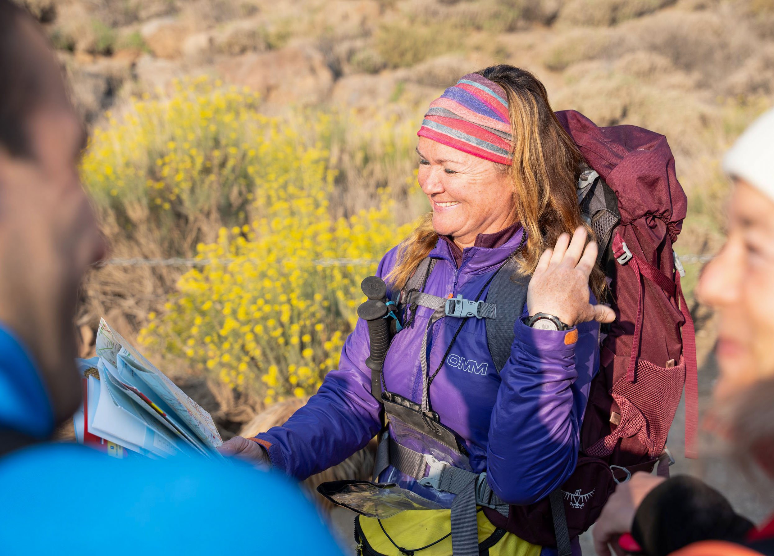 A smiling Leanne Rive with a colorful headband and purple jacket holding a map while on a hiking trail in a desert landscape, surrounded by two other hikers.