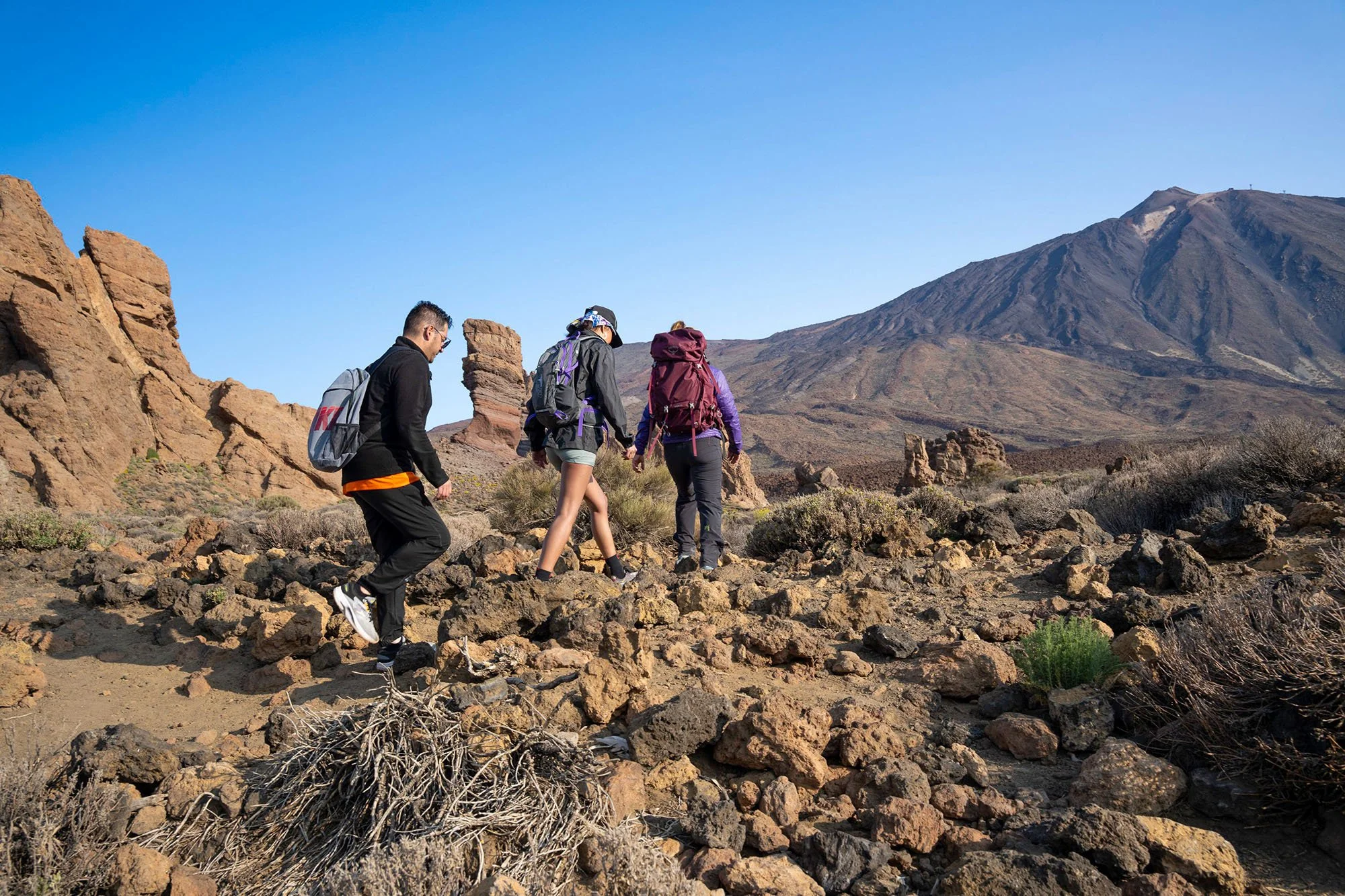 Three hikers with backpacks walking on rocky terrain in a desert landscape with mountains in the background under a clear blue sky.